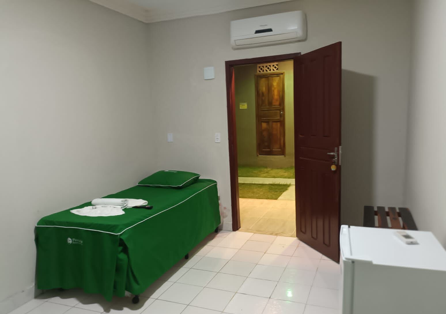 A small room with a green-covered massage table, a white air conditioning unit, and a wooden door leading outside.