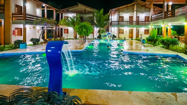 A swimming pool with a waterfall feature, surrounded by buildings with outdoor seating and greenery. - Pousada Recanto do Mar (Camocim)