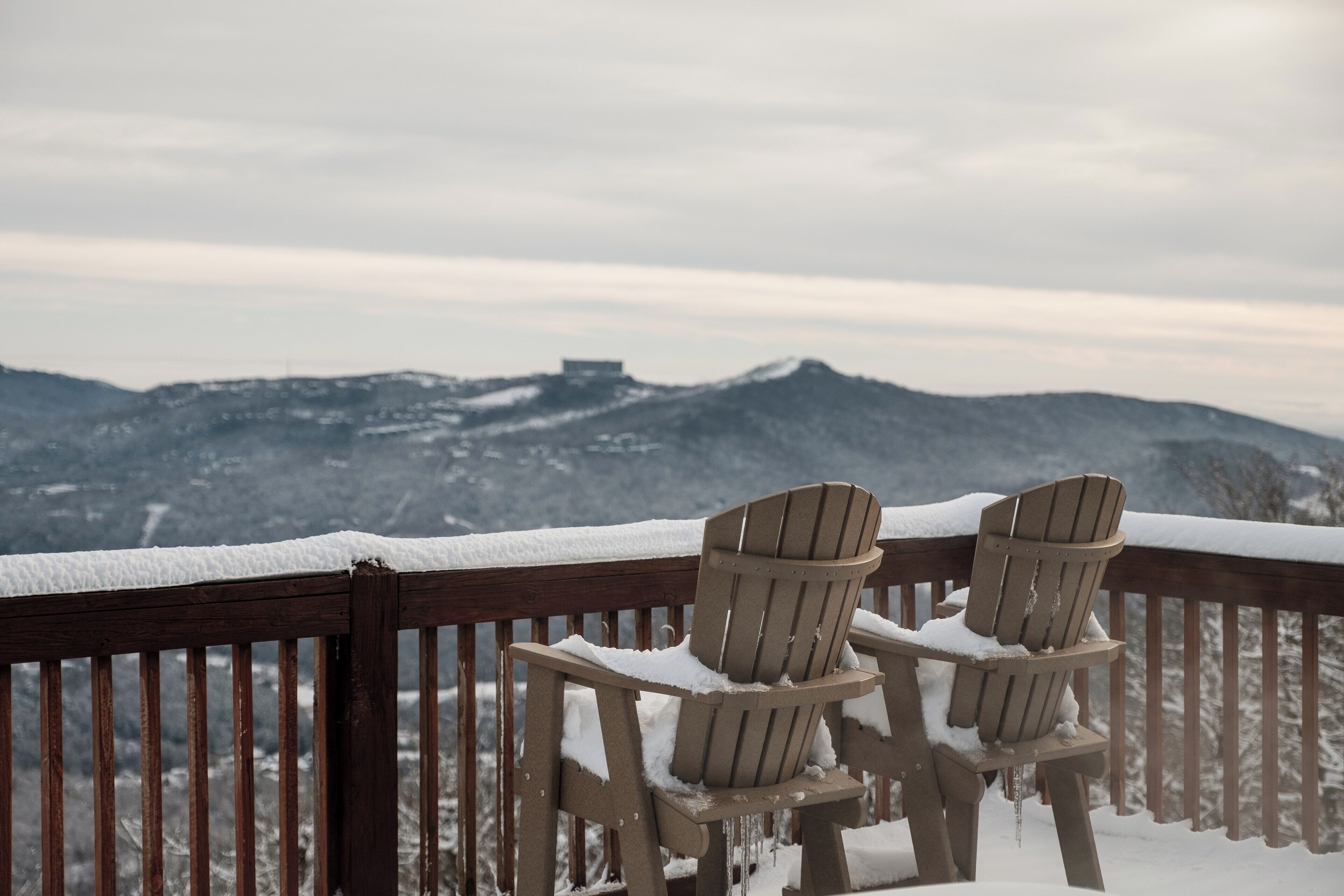 Two wooden Adirondack chairs on a snow-covered deck with a view of a mountainous landscape.