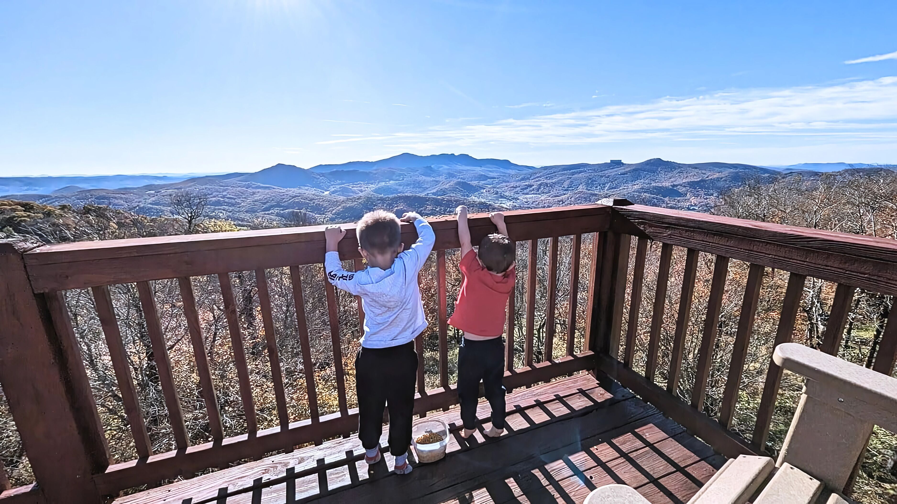 Deux enfants se tiennent sur une terrasse en bois, d’où l’on peut admirer des montagnes et un ciel dégagé.