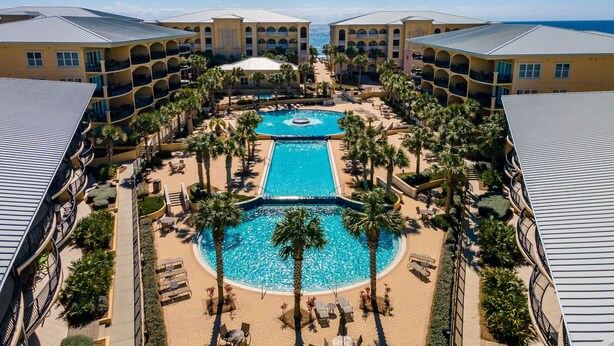 Un resort con piscina central rodeada de palmeras y tumbonas, junto a un edificio con balcones.