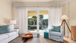 Una sala de estar moderna con sofá, sillón, mesa de centro, lámpara y vistas a un espacio exterior con palmeras a través de amplias puertas de cristal.