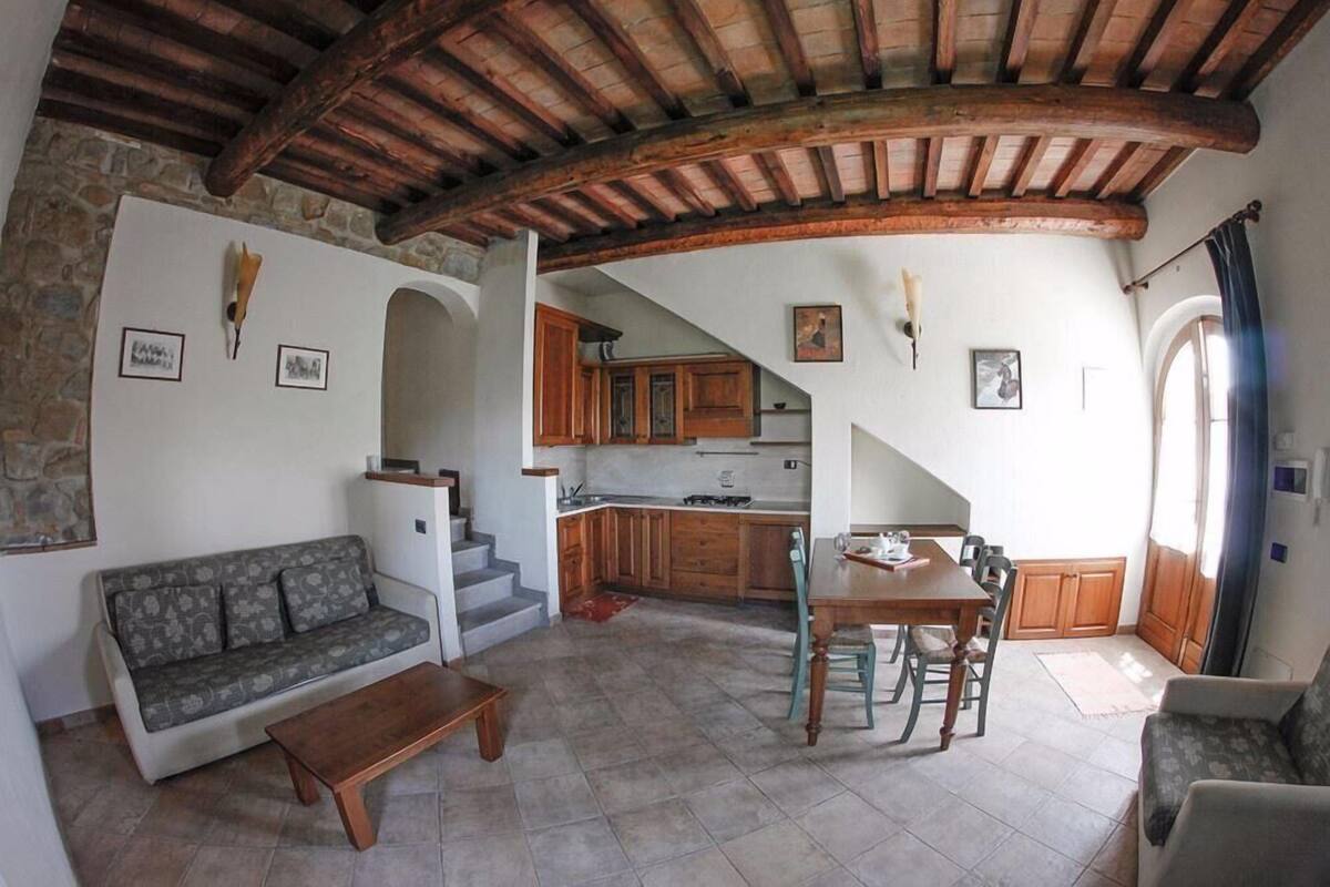 A cozy living area with a sofa, coffee table, and dining table. There is a kitchen in the background with wooden cabinets and a staircase leading to another level.