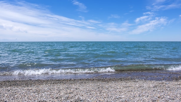 A pebbled beach with clear blue water and a calm sea under a partly cloudy sky. - Lake Erie House Near Cedar Point & The Beach (Huron)