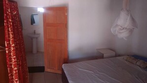 A bedroom with a bed, a bedside table, a hanging laundry rack, a wooden door, and a small window.