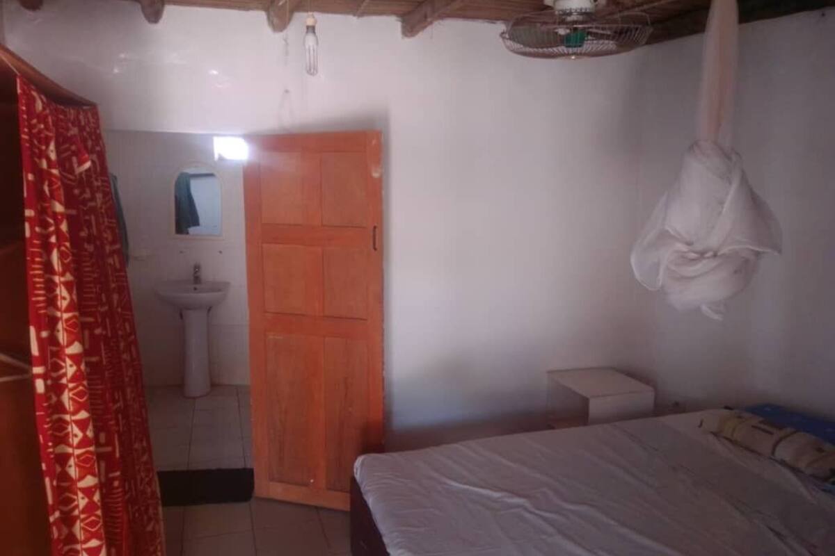 Une chambre à coucher comprenant un lit, une table de chevet, un étendoir suspendu, une porte en bois et une petite fenêtre.