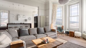 A spacious living room with a grey sofa, wooden coffee table, and a view of the outdoors through large windows. - 5* luxury pet friendly house in Woolacombe with hot tub and sea views (Woolacombe)