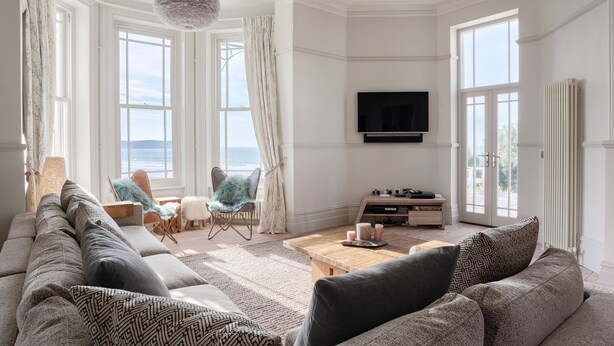 A spacious living room with a large sofa, a coffee table, and a television mounted on the wall. There are large windows providing a view of the outdoors. - 5* luxury pet friendly house in Woolacombe with hot tub and sea views (Woolacombe)