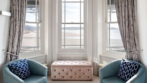 A room with a bay window overlooking a beach, featuring two upholstered armchairs and a tufted ottoman. - 5* luxury pet friendly house in Woolacombe with hot tub and sea views (Woolacombe)