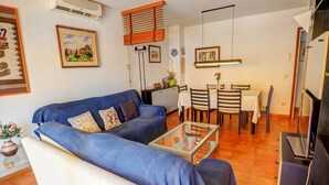 A living room with a blue sofa, a glass coffee table, a dining area with a table and chairs, and a TV on a stand. - Oasis Sinia by Hello Homes Sitges (Sitges)