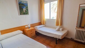 A hotel room with two beds, a wooden nightstand, a small table, a radiator, and a window with curtains. - Oasis Sinia by Hello Homes Sitges (Sitges)