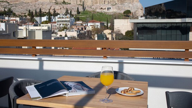 Ein Tisch mit einem Buch, einem Glas Orangensaft und einem Tablett Gebäck, mit Blick auf eine Stadt und antike Ruinen im Hintergrund.