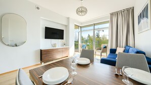 Un espace repas moderne avec une table en bois dressée pour quatre personnes, un téléviseur à écran plat et une grande fenêtre agrémentée de rideaux.