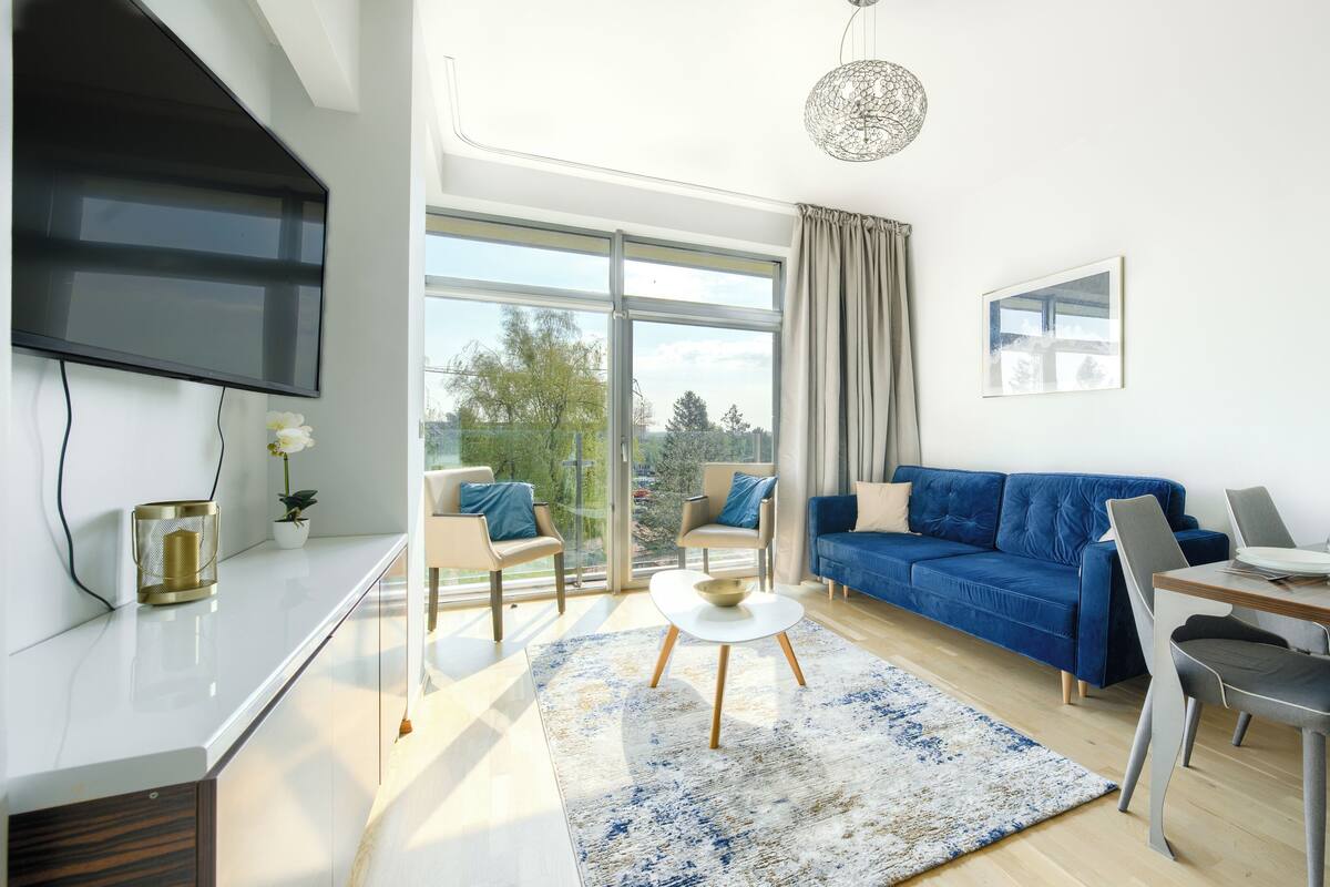 A modern living room with a blue sofa, a dining table with chairs, a TV mounted on the wall, and a large window with curtains.
