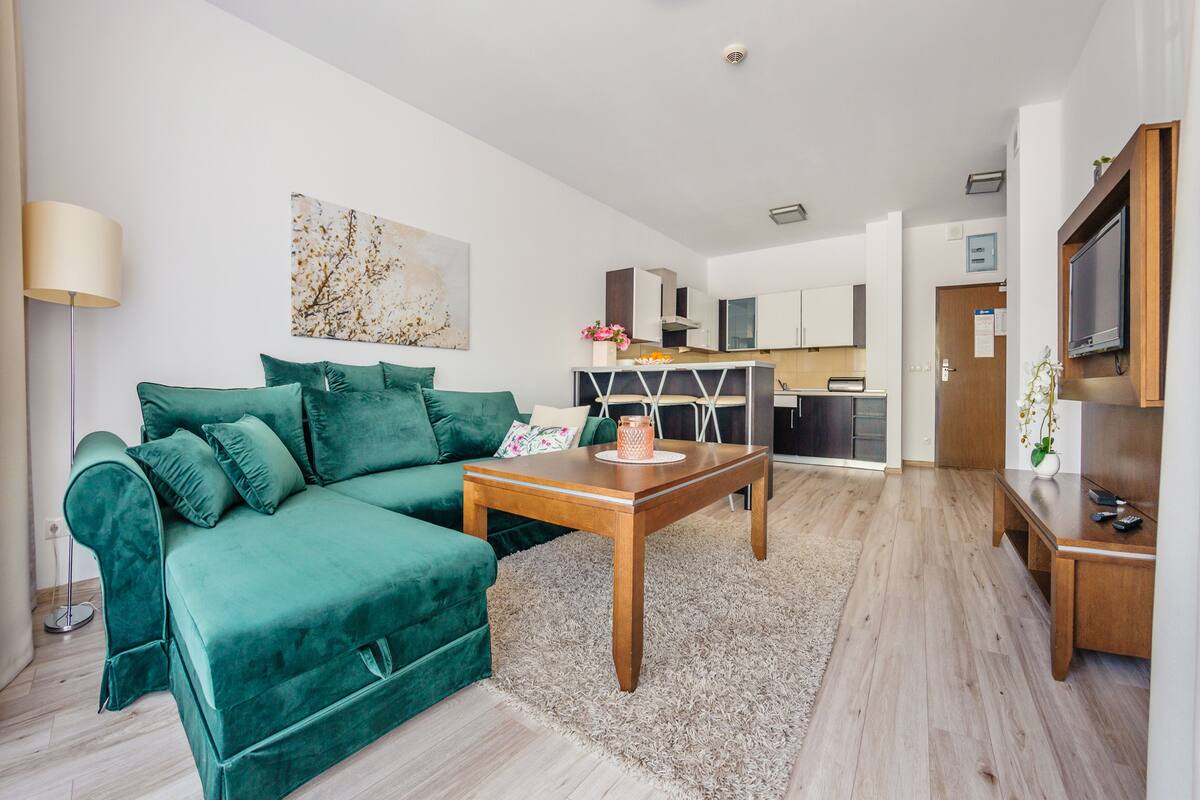 A modern living room with a green sofa, a wooden coffee table, and a kitchen area in the background.