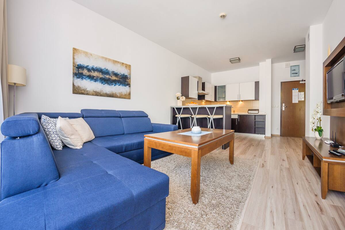 A modern living room with a blue sofa, a wooden coffee table, and a kitchen in the background.