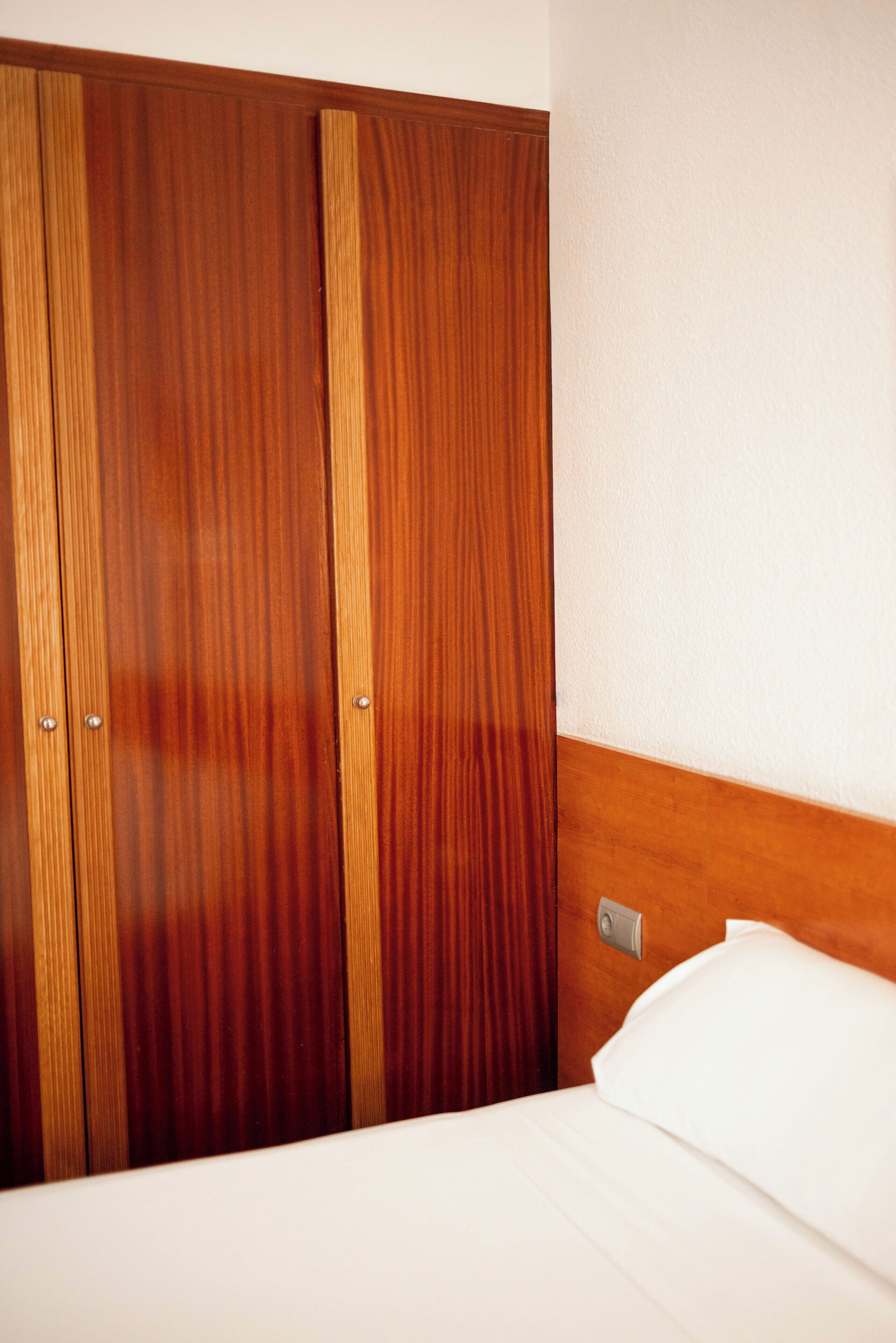 A hotel room with a wooden wardrobe, a bed with white bedding, and a wall-mounted light switch.