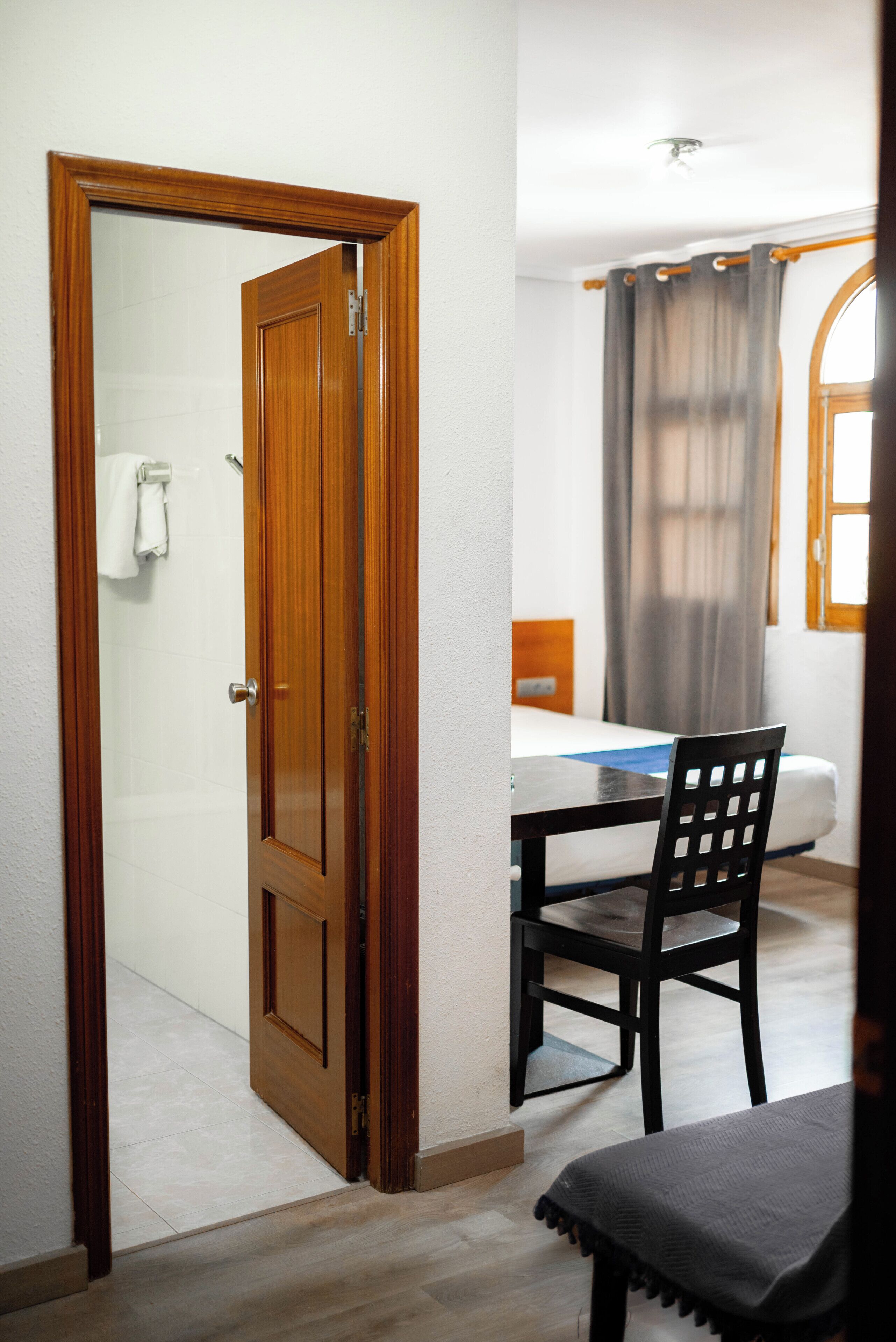 A room with a wooden door, a bed with a headboard, a table with a chair, and a window with curtains.