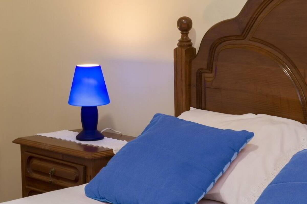 A bed with white linens and blue pillows, a wooden headboard, and a nightstand with a blue lamp.