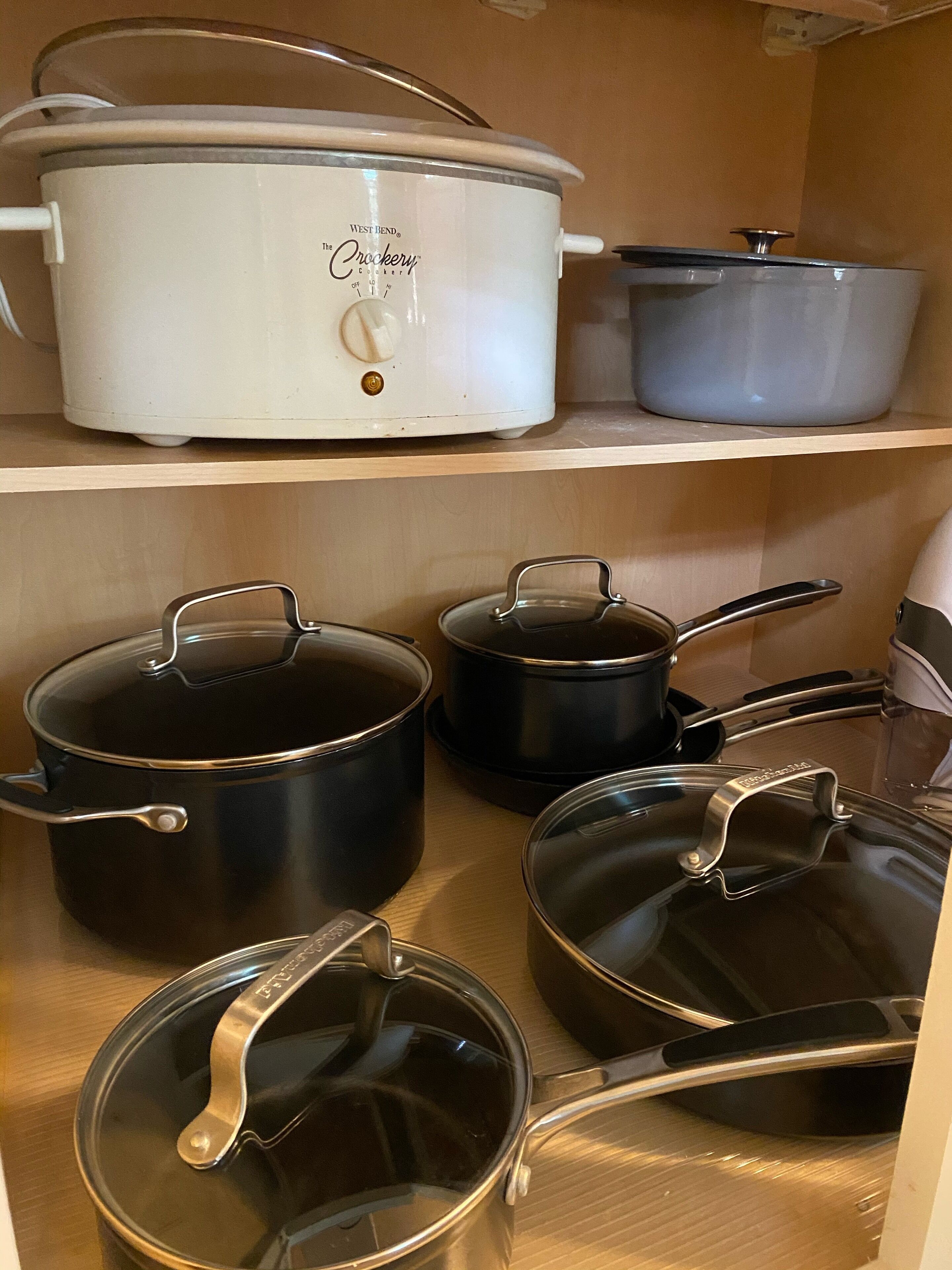 Une étagère de cuisine contenant divers ustensiles de cuisine, notamment un autocuiseur West Bend Crockery blanc et plusieurs casseroles noires avec couvercles.