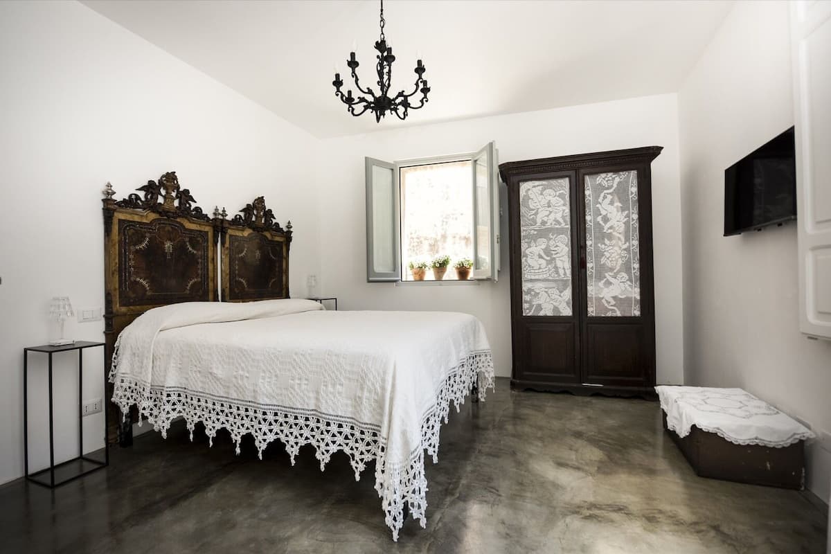 Un dormitorio con una cama grande, un armario de madera, una araña y una ventana con cortinas de encaje.