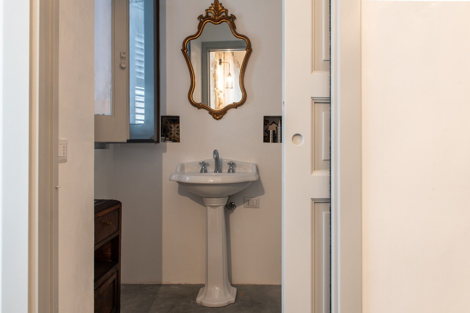 Een badkamer met een wastafel op een voetstuk, een spiegel met een sierlijk omlijsting, en een deur naar een andere kamer.