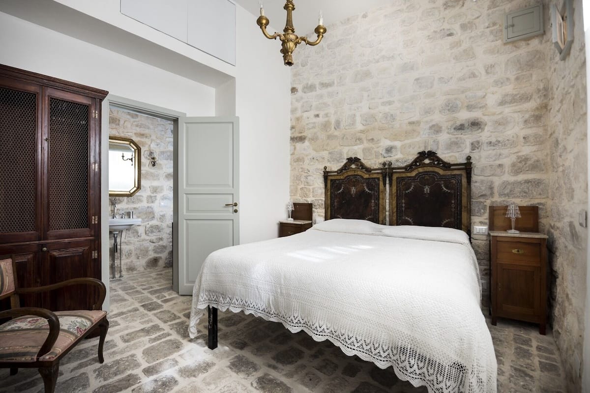 Un dormitorio con pared de piedra, cama con cubierta blanca de encaje, mesita de noche de madera, silla de madera y una araña.