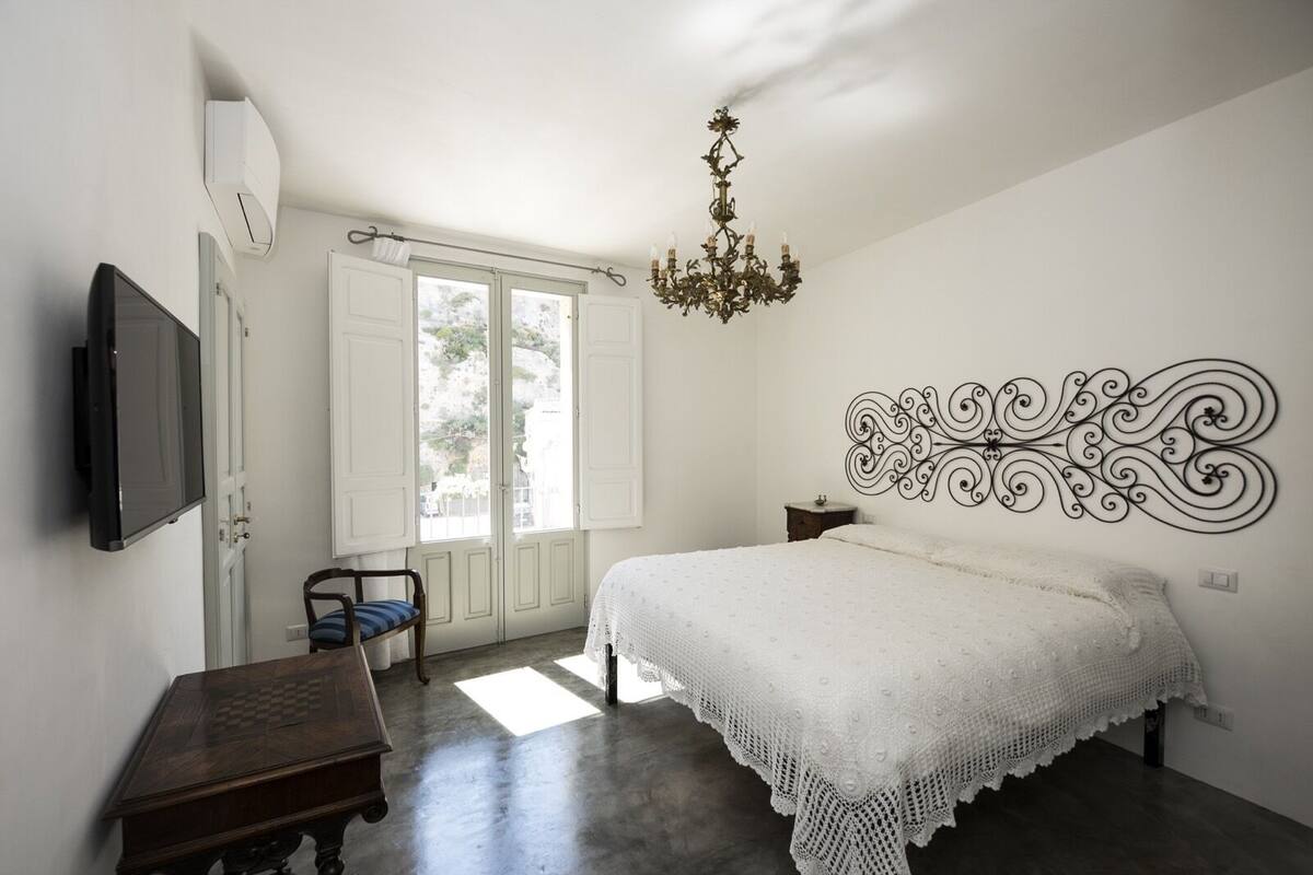 Un dormitorio con cama, televisor, mesa de madera, silla, ventana con cortinas y candelabro.