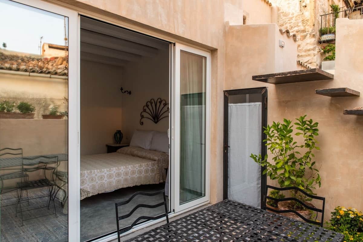 Un dormitorio con cama, mesita de noche, un adorno en la pared, una puerta corrediza de vidrio y un balcón pequeño con mesa y sillas.