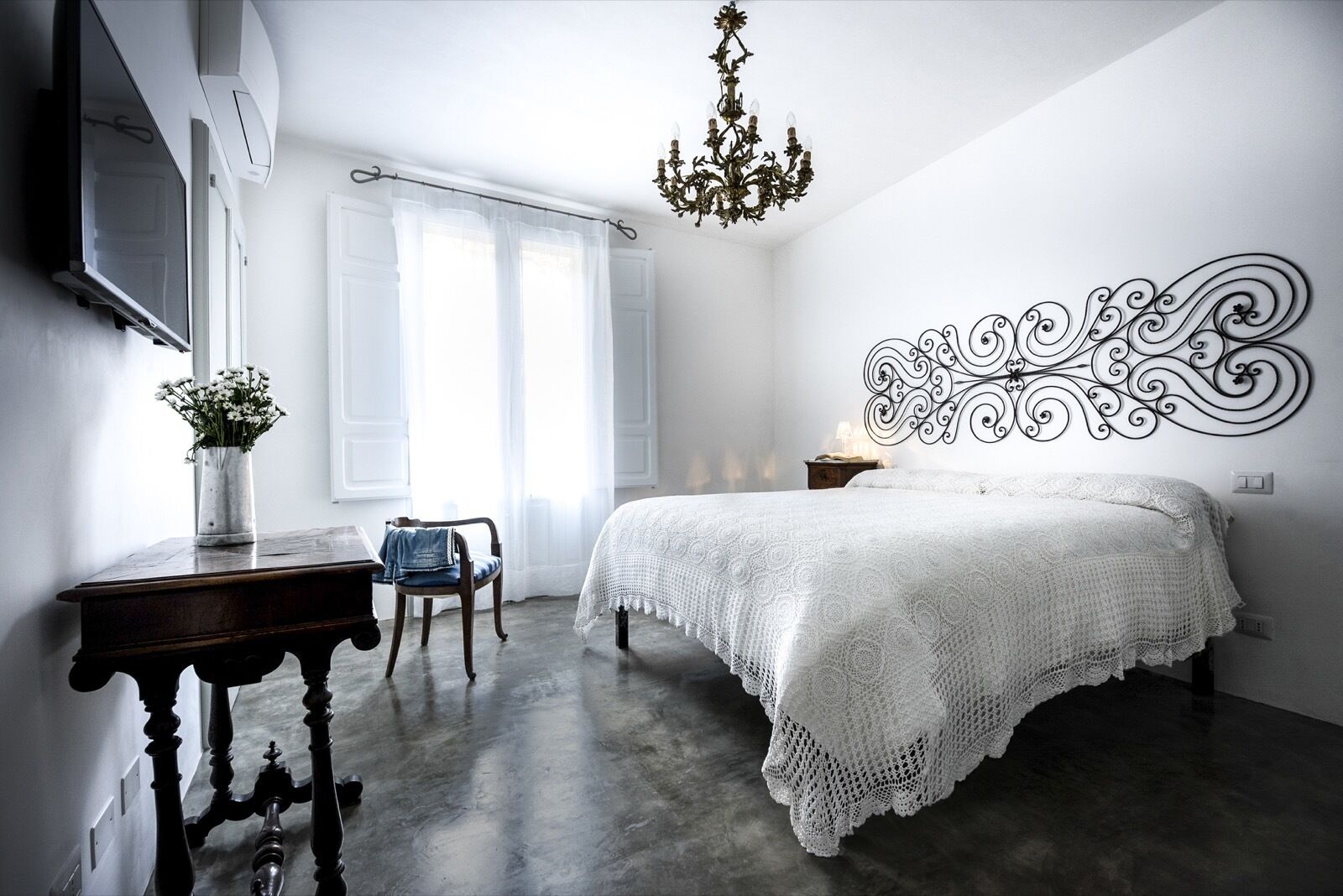 A bedroom with a large bed, a chandelier, a decorative wall piece, a desk with a vase of flowers, and a chair.