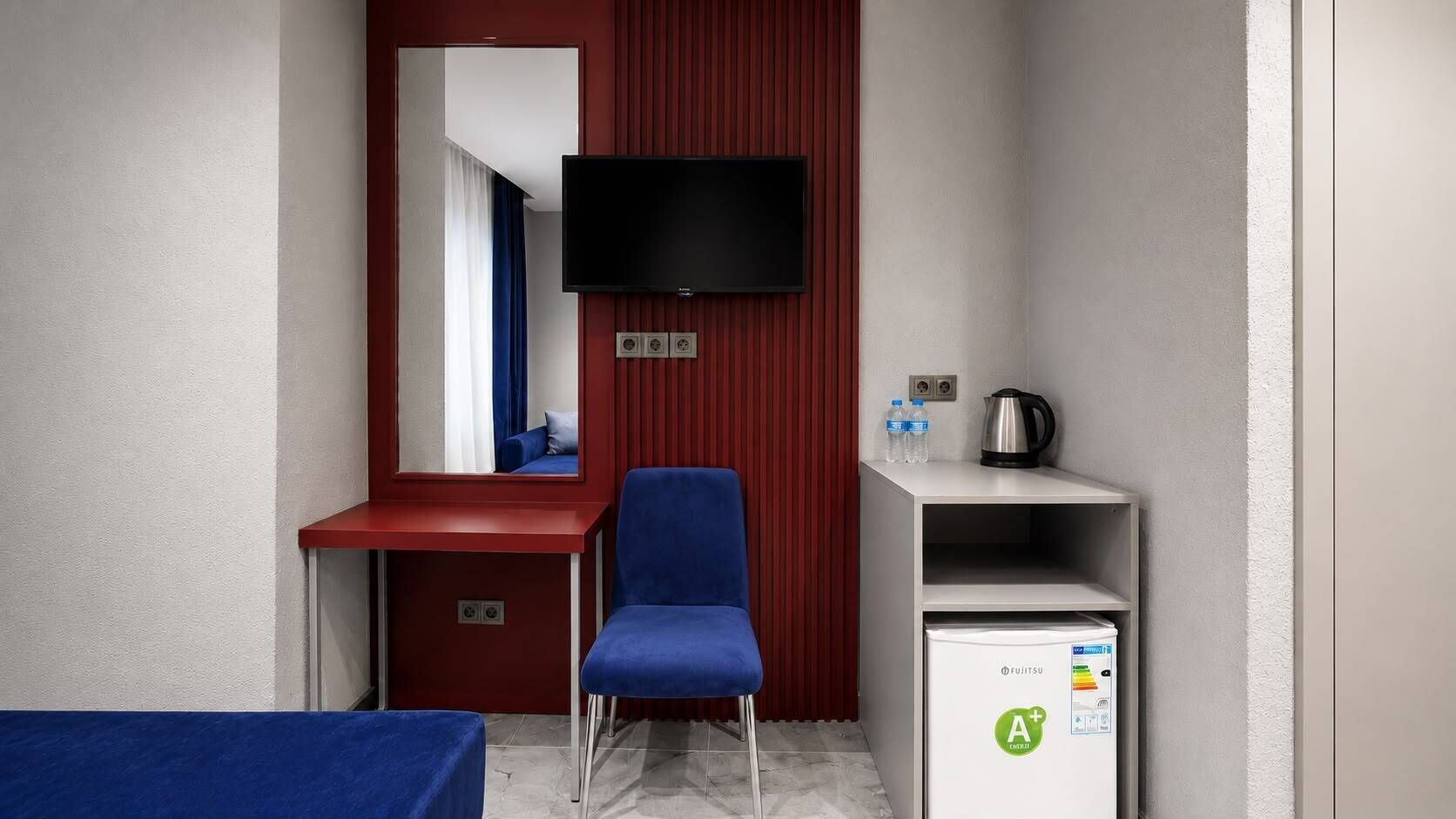 A small hotel room with a single blue chair, a television mounted on the wall, a mini-fridge, and a kettle on a shelf.
