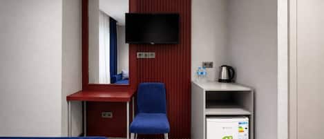 A small hotel room with a single blue chair, a television mounted on the wall, a mini-fridge, and a kettle on a shelf.