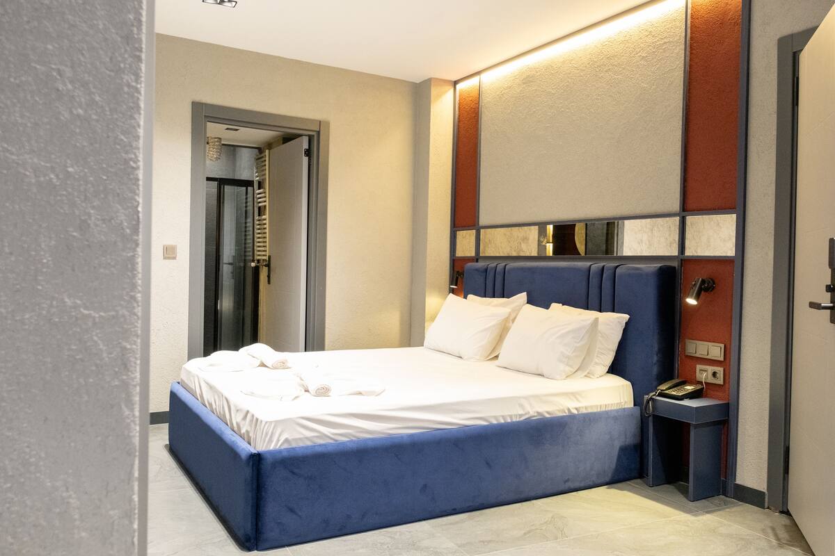 A modern hotel room with a large bed, a bedside table with a phone, and a door leading to another room.