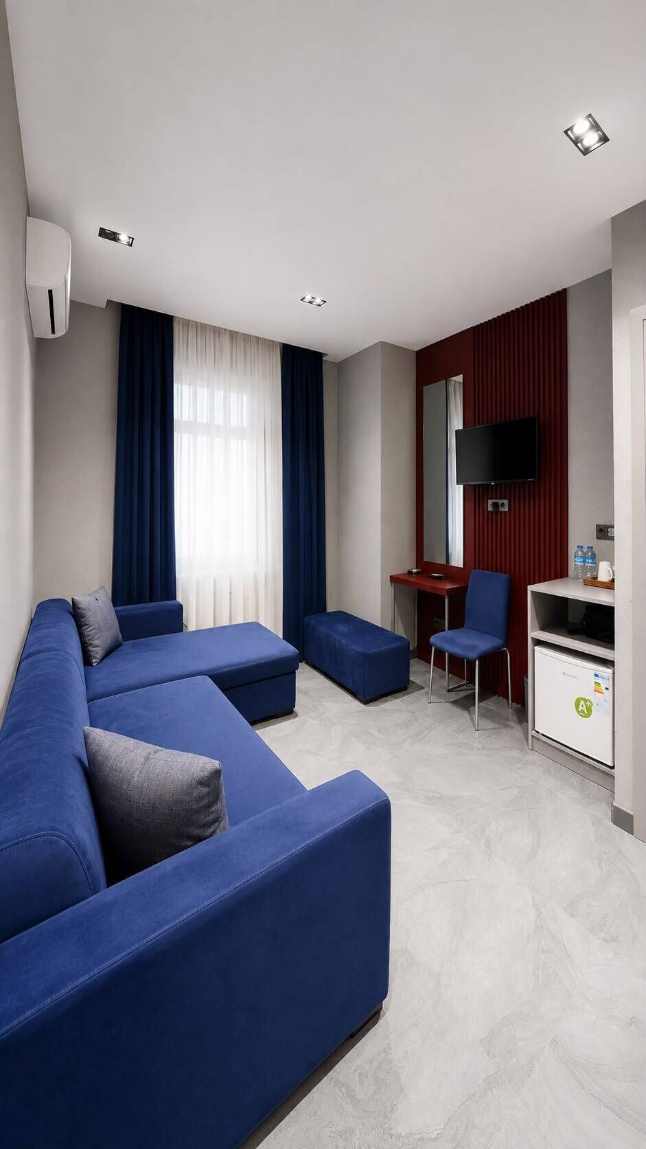 Una habitación de hotel moderna con un conjunto de sofá azul, un escritorio con una silla, un televisor montado en la pared y un minibar.