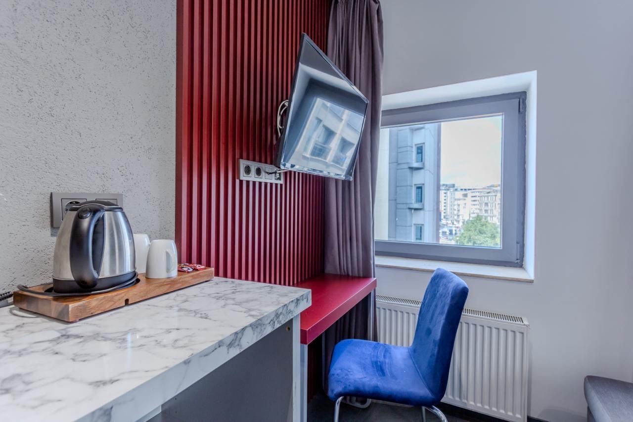 Una habitación de hotel pequeña con una pared de acento roja, un televisor de pantalla plana montado en ella, una encimera de mármol, una silla azul y una ventana con vista a la ciudad.