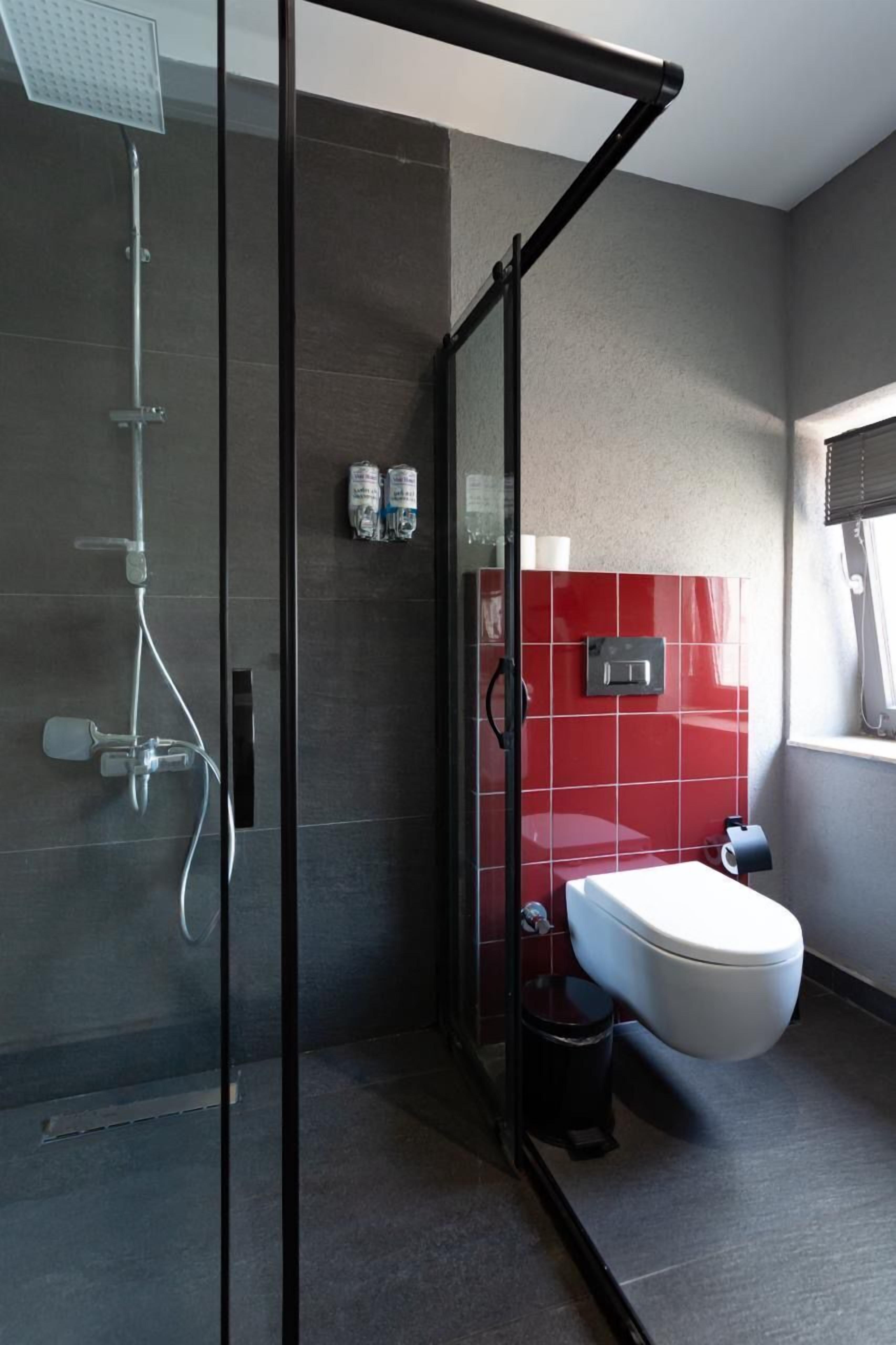 Une salle de bain moderne avec une douche en verre, un mur recouvert de carreaux rouges, un lavabo blanc et une poubelle noire.