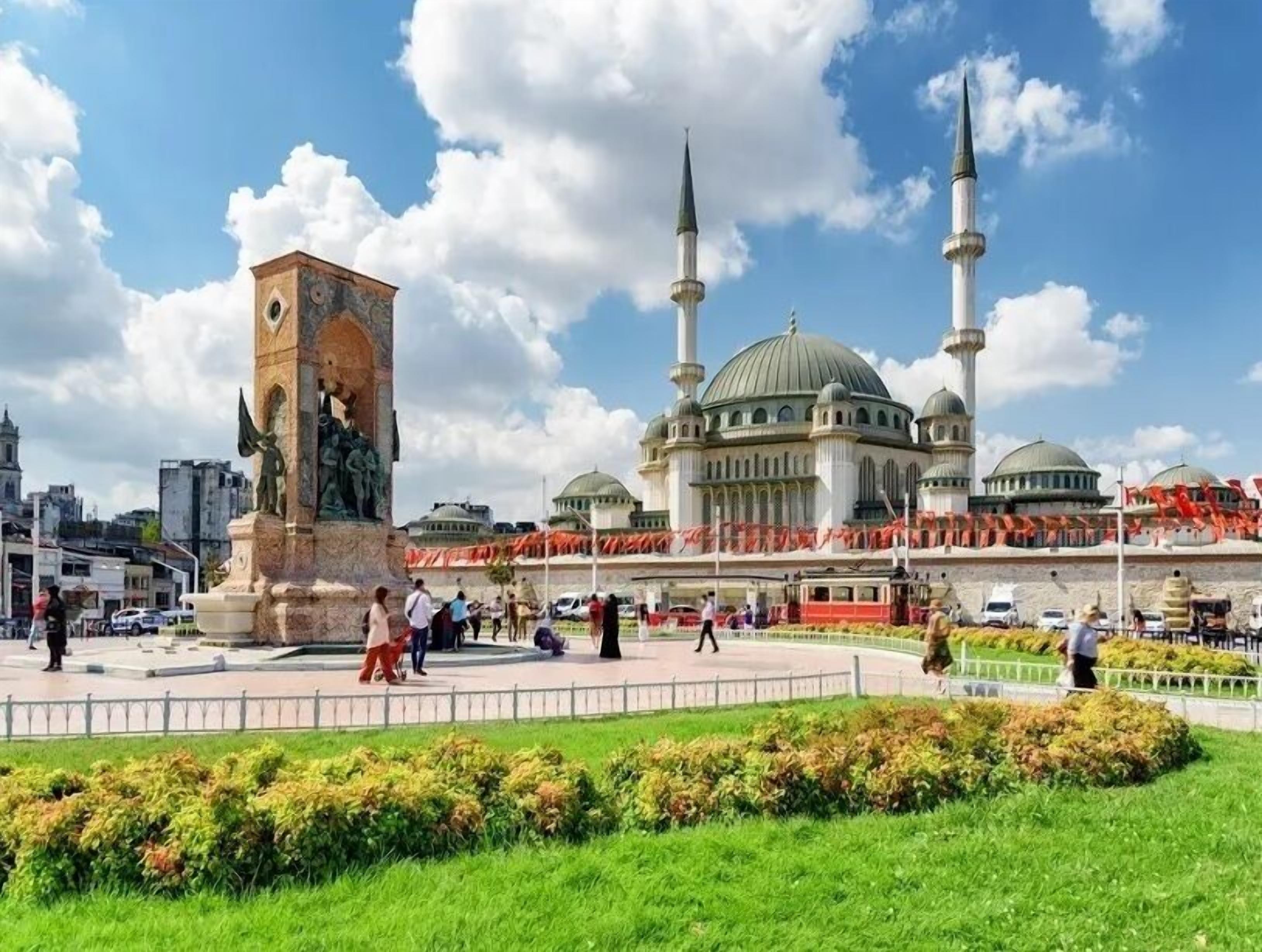 Una mezquita con dos minaretes y una cúpula grande, rodeada por una plaza pública con una estatua y zonas verdes.
