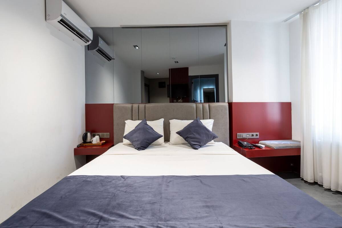 A modern hotel room with a large bed, a bedside table with a phone and a lamp, and a view of the hallway.