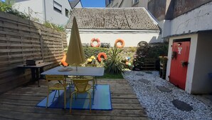 Een terras met een tafel en stoelen, omringd door een houten hek, een rode deur en een tuin met diverse planten en decoratieve objecten.