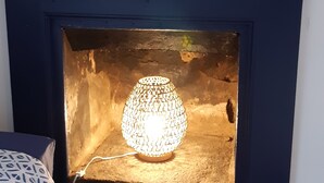 Een open haard met een decoratieve lamp, daarboven twee ingelijste foto's en een gestreept kussen op de vloer.