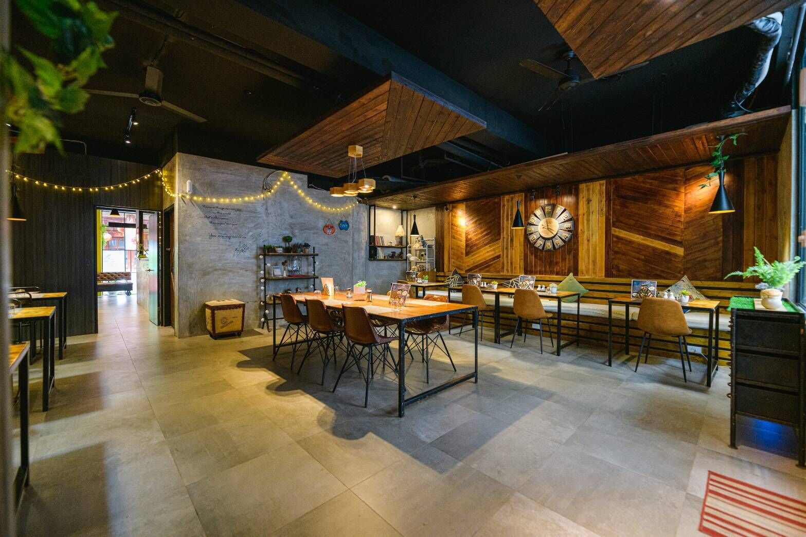 A modern restaurant with exposed wooden beams, a central dining area with tables and chairs, a bar area, and a large wall clock.