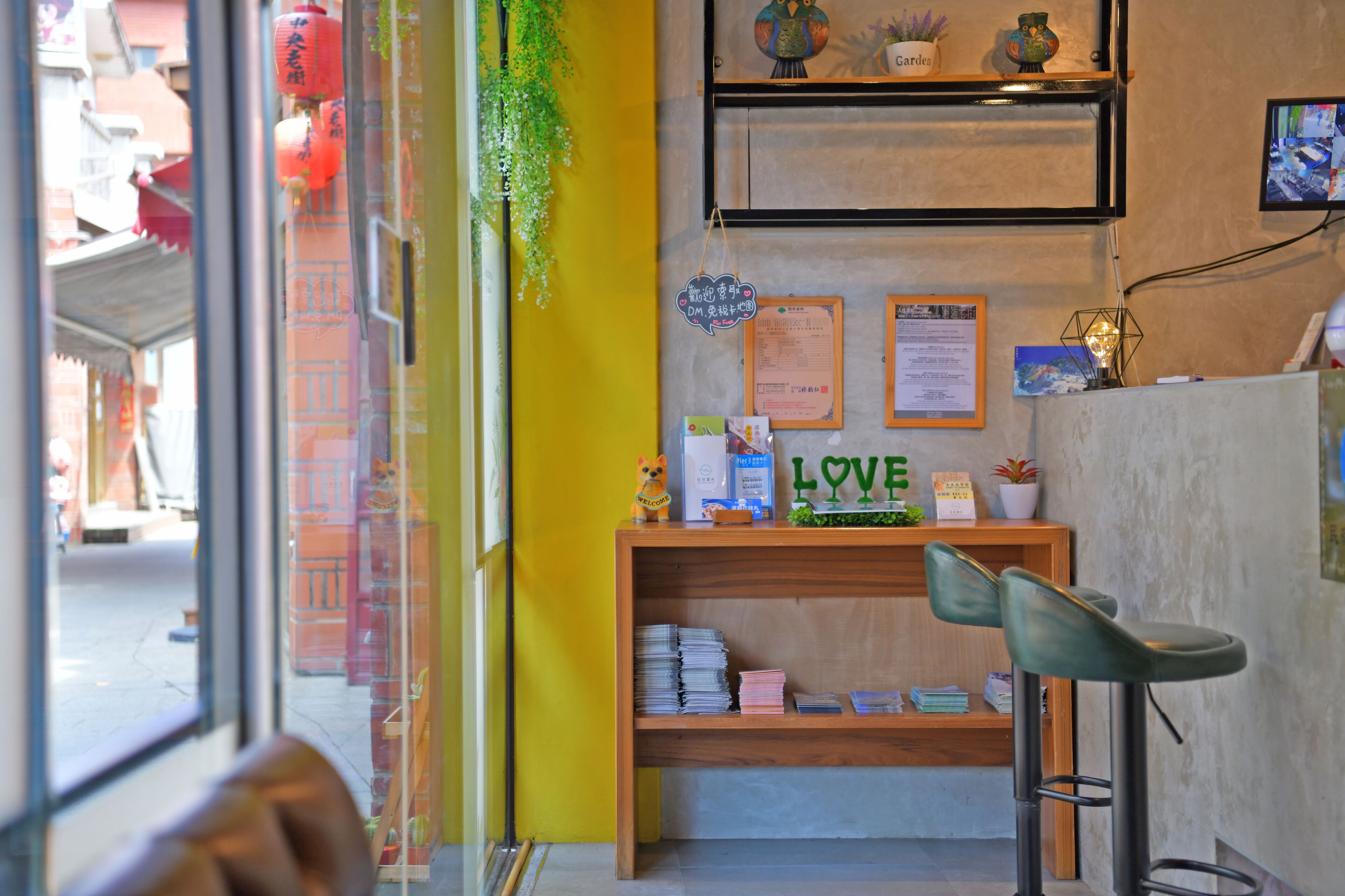 A bar area with a wooden shelf displaying various items, a bar stool, and a window with a view of the street.