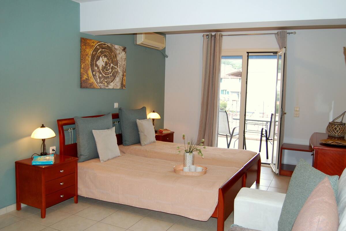 A double bed with a beige bedspread, two bedside tables with lamps, a small table with a vase and flowers, and a view of the outside through a sliding door.