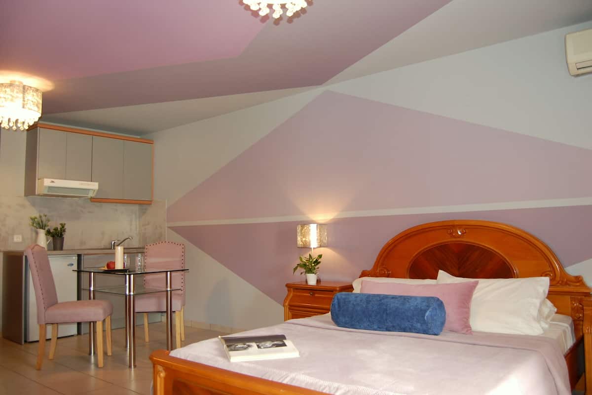 A bedroom with a bed, bedside table, lamp, and a small kitchenette area with a chair and table.