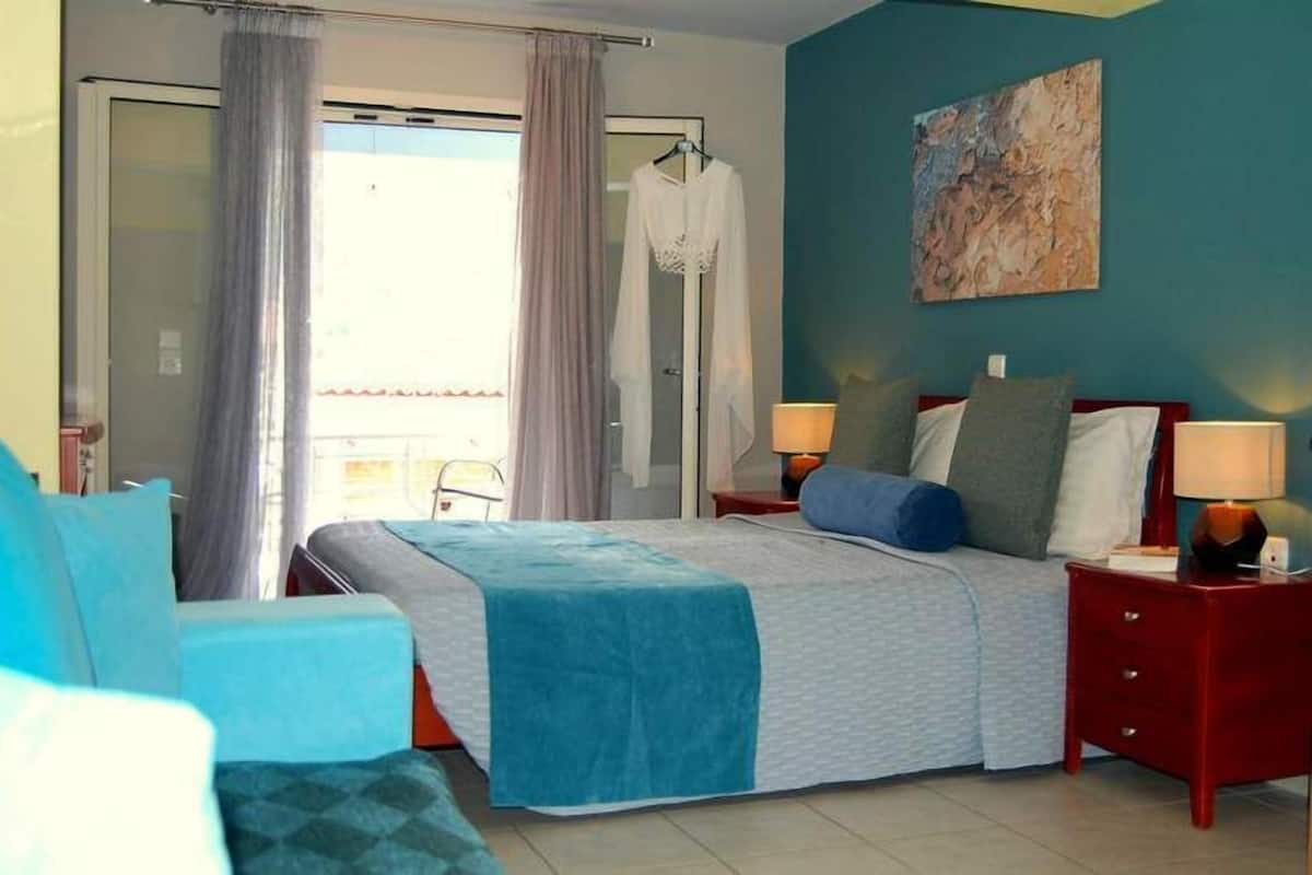 A hotel room with a bed, two bedside tables with lamps, a chair, a balcony with a view, and a painting on the wall.