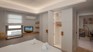 A modern hotel room with a large bed, a television, a wardrobe, and a small seating area with a table and chairs. - Acropolis Golden Suites | 3 by GHH (Athina)