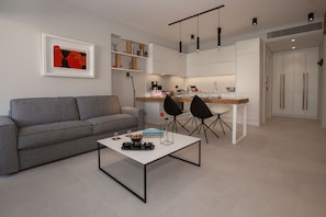 Un salón moderno con un sofá gris, una mesa de centro blanca y sillas negras. Se observa una zona de cocina con armarios blancos y una encimera de madera.