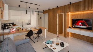 A modern living room with a grey sofa, a coffee table with books and a candle, a TV mounted on a wooden wall, and a kitchen area with white cabinets and a wooden countertop. - Acropolis Golden Suites | 3 by GHH (Athina)