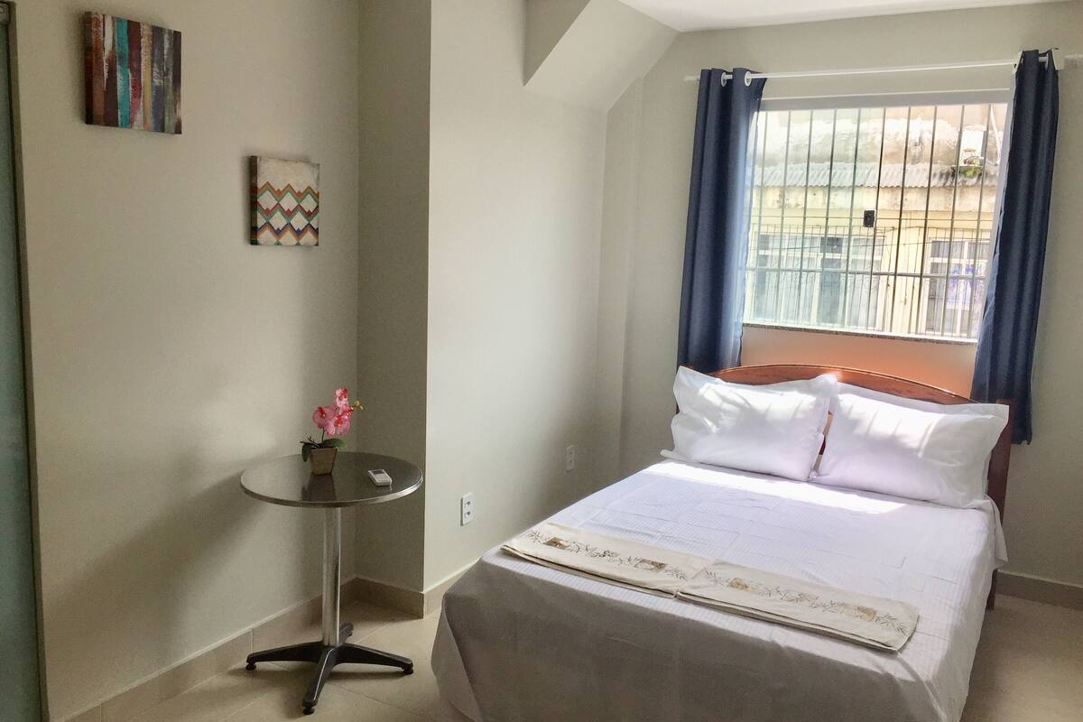 Ein ordentlich eingerichtetes Schlafzimmer mit einem Bett, einem kleinen runden Tisch mit einer Topfpflanze und einem Fenster mit Vorhängen.