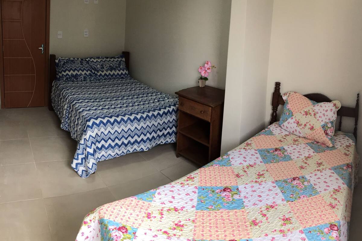 Ein Schlafzimmer mit zwei Betten, eines mit einer blau-weißen Bettdecke und das andere mit einer Blumensteppdecke. Dazwischen stehen die Betten an einem Nachttisch mit einer Vase mit rosa Blumen.